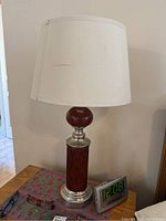 Lamp on wooden table next to digital clock and glasses. Lamp has wooden grooved column base, silver metal accents, and cream drum shade.