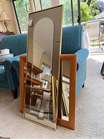 Three mirrors leaning against a blue chair and a leopard patterned chair. The tallest mirror is rectangular with an arched top and a thin gold-tone metal frame. One smaller rectangular mirror with a similar gold-tone metal frame and another with a wider wooden frame.