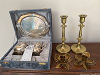 Silver plate creamer, sugar bowl, serving tray in fitted blue velvet case and two brass candlesticks with two brass flower-shaped candle holders on a wooden surface.