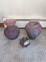 Three handcrafted coloured glass vases arranged on a carpet in front of a baseboard heater. Two larger vases and one smaller decorative bowl-like piece.