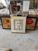 Three framed wall art pieces arranged in front of a plaid couch: largest rectangular abstract art in neutral tones; two smaller floral prints with orange and red flowers; framed textured 3D abstract piece with pastel colors in gold frame.