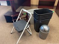 Five black plastic trash bins stacked, small silver dome-lid desk trash bin, white step stool with black tread, wooden magazine rack and file organizer in background.