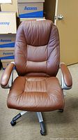 Front view of the brown padded office chair showing seat, backrest, and armrests with silver metal frame and black caster wheels.
