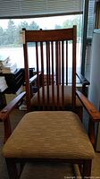 Front-facing photo of one wooden chair with vertical slat back and beige upholstered seat, placed indoors near window with blinds.