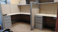 Two joined L-shaped office cubicle desks with fabric partitions, metal filing cabinets, and central divider visible.
