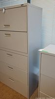 Set of five drawers in a tall gray metal cabinet, showing the full front and side profile under natural light next to a window.