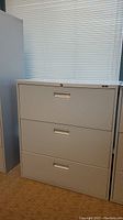 Front view of light grey metal filing cabinet showing three drawers and lock on top drawer.