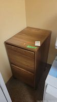 Front and side view of a 2 drawer filing cabinet with a wood-style finish in an office corner.