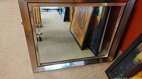 Clear rectangular mirror with a wooden frame showing the reflection of carpeted floor and nearby paintings.