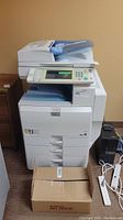 Full front-right view of Ricoh Aficio MP 4001 printer showing paper trays, document feeder, control panel, cardboard box with toner cartridges, UPS and cables.