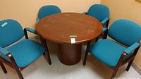 Photo showing round brown wood table with cylindrical pedestal base surrounded by four blue upholstered chairs with wood frames and armrests, room corner background.