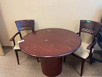 Wide view of round wooden table with two captains chairs placed on either side against a plain wall, showing overall condition and design.
