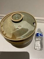 Top view of the lidded pottery bowl with a water bottle beside it for scale. The lid is glazed in two tones with a knob handle and a notch.