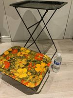 Front view of two folding trays: a colorful floral patterned tray in front folded down and a taller black metal tray with folding legs at the back.