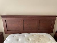 Photo shows a wooden headboard attached to a full size bed frame with part of a mattress visible. The headboard has three recessed panels with a crown molding top and a natural wood finish.