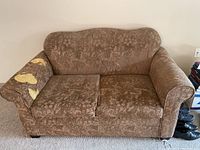 Front view of brown loveseat showing floral pattern upholstery with visible damage on left armrest.