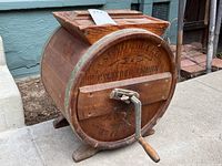 Photo showing the full wooden cylinder butter churn with crank handle on the front and wooden base supports, placed outdoors on a sidewalk.