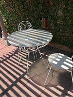 Full view of the white iron bistro set with round glass top table and two matching chairs outdoors on stone patio with surrounding green ivy wall.