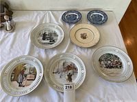 Overview of six hanging decorative plates laid on a table, showing various Carl Larsson scenes and two smaller blue patterned plates.
