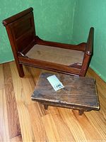 Wooden doll or small pet bed frame with paneled headboard and footboard next to a small wooden step stool, placed on wood floor in corner of green textured wall room.