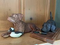 Two ceramic hunting dog figurines, one brown with a duck, one black with binoculars on a wooden base, displayed on wood shelf.
