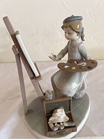 Front and side view of the porcelain figurine depicting a girl painting at an easel, showing palette and paintbox detail.
