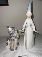 Two Lladro figurines, clown with dog on dice and fairy godmother in white gown, shown standing together on a white surface against a beige wall.