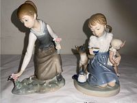 Two Lladro figurines displayed side by side. One depicts a girl picking flowers, the other a girl holding a child doll with a cat beside her.