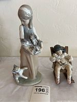 Photo showing two Lladro porcelain figurines: girl holding cats and girl sleeping in rocking chair holding doll, with measuring note visible.