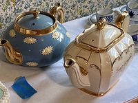 Both teapots shown together to show size difference and style contrast: one round blue with cream flowers and gold, one rectangular cream with gold detail.