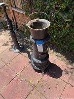 Full view of vintage metal parlor kerosene oil heater with metal pot on top, placed outside on pavers with a green shrub background.