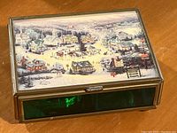 Photo of the music box with a winter village Christmas scene on the lid, showing metal and green tinted glass sides.