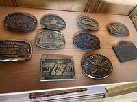 Collection of ten vintage brass belt buckles displayed on a wooden surface, showing front faces with embossed shooting and hunting designs, logos and inscriptions.