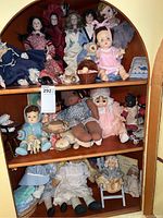 Wide view of three shelves within a wooden corner unit displaying a collection of vintage dolls of various sizes, styles, and clothing, including porcelain and cloth dolls.
