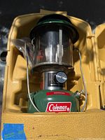 Front view of the green-top Coleman lantern in the yellow carrying case, showing the Coleman label and valve wheel.
