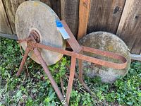 Full view of the vintage sharpening wheel showing two large circular grindstones on rusted iron tripod frame in overgrown foliage.