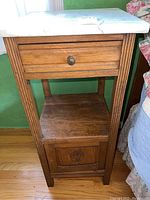 Front view of vintage wood side table with white marble top, one drawer with round metal pull, open middle shelf, and lower cabinet door with carved detail.