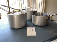 Three All-Clad stainless steel MasterChef pots with two pots having lids placed on a table with natural light from window in background.