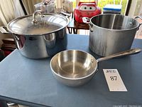 Top view of the All Clad pots and pan showing the large 11 inch stock pot with lid, the smaller 8 inch stock pot without lid, and a small 6.5 inch pan with handle on a blue surface.