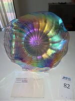 Front view of the circular blown glass plate showing rainbow iridescence and spiral pattern on a clear stand with label for Nowak Glass Studio and piece number 1226.