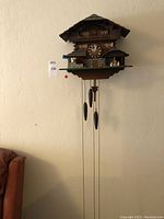 Front view of chalet-style wood cuckoo clock mounted on wall with three pine cone weights and chains hanging beneath.