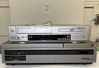 Sylvania LaserDisc player stacked below GO Video VHS/DVD combo player with all front controls visible.