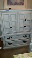 Front view of painted wood armoire dresser showing upper paneled cabinet doors with round brass knobs and two lower drawers with brass handles.