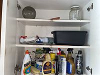 Photo of open white cabinet shelves showing multiple cleaning product bottles, black basin, and glass jar