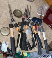 Photo showing multiple gardening hand tools with wooden handles and metal blades, including hedge clippers, snips, and a hammer, laid out on a carpeted surface with two small round metal tins.