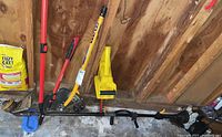 Garden tools including a long-handled edger, trimmer, and yellow blade attachment laid horizontally on a concrete surface in a wooden garage.
