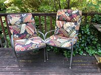 Pair of metal patio chairs with colorful leaf patterned cushions set outdoors on a wooden deck, showing the front and side views.