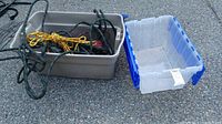 Large light gray plastic storage bin containing coiled ropes and cords, and empty translucent plastic bin with blue handles next to it on pavement