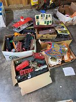 View of boxes containing black locomotives and various train cars in two cardboard boxes. Surrounding train village buildings and boxes of accessories visible.