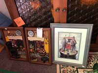 Two smaller framed rustic kitchen interior dioramas depicting a fireplace and stove scene, and a larger framed watercolor print showing two men on a boat.